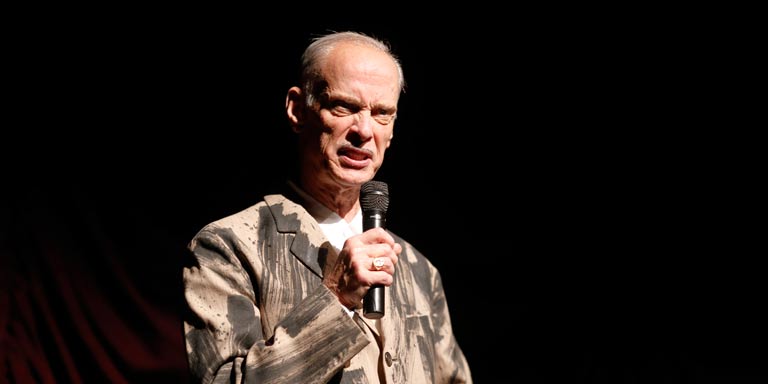 John Waters speaking on a microphone on stage